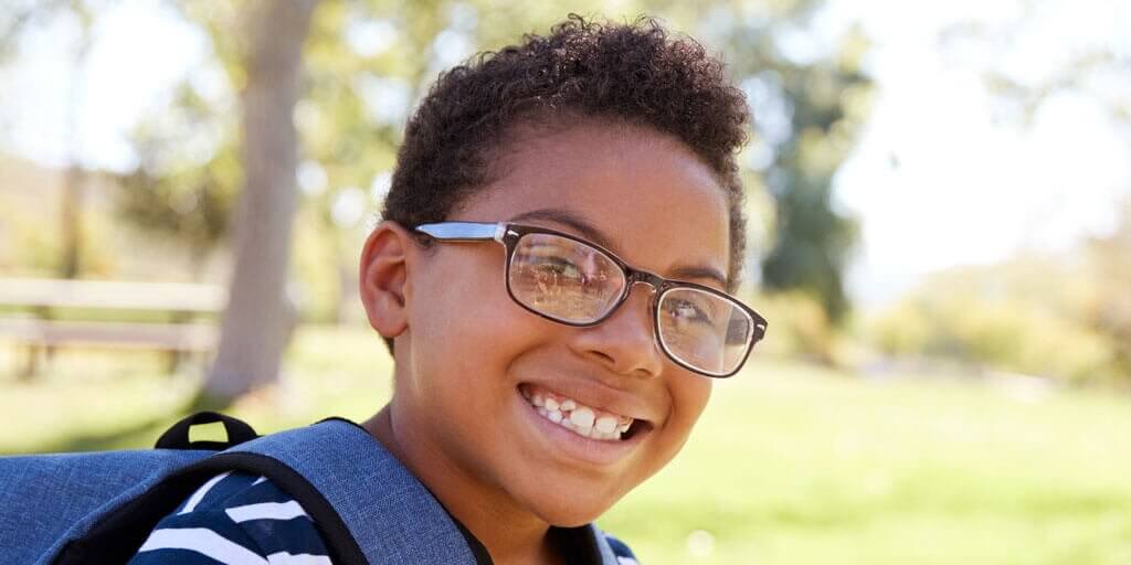 what-is-strabismus-1024x512 A young boy with eyeglasses and a backpack is smiling outdoors.